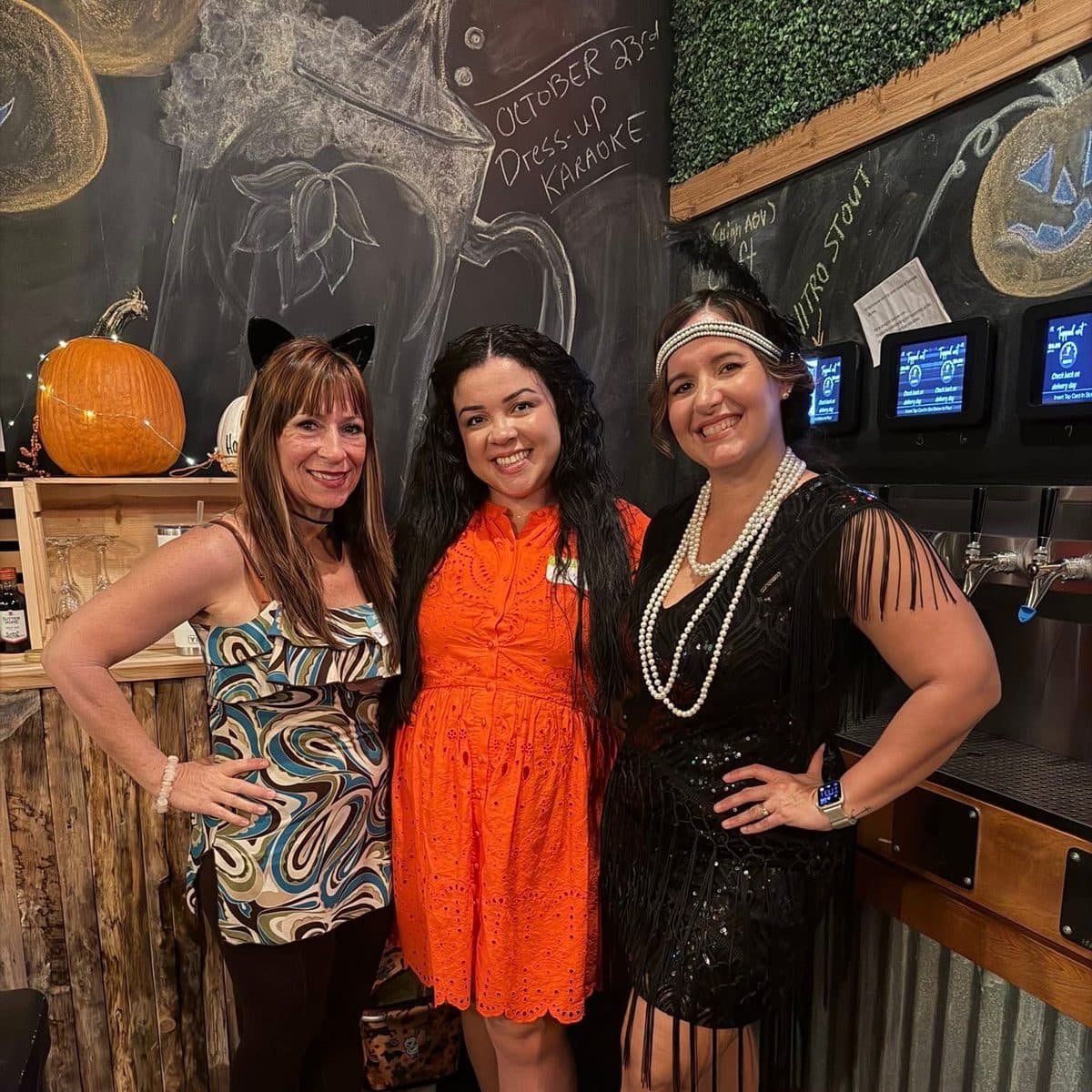 Nelly in an orange dress with a woman in cat ears and another in a flapper costume, beside the self-pour taps
