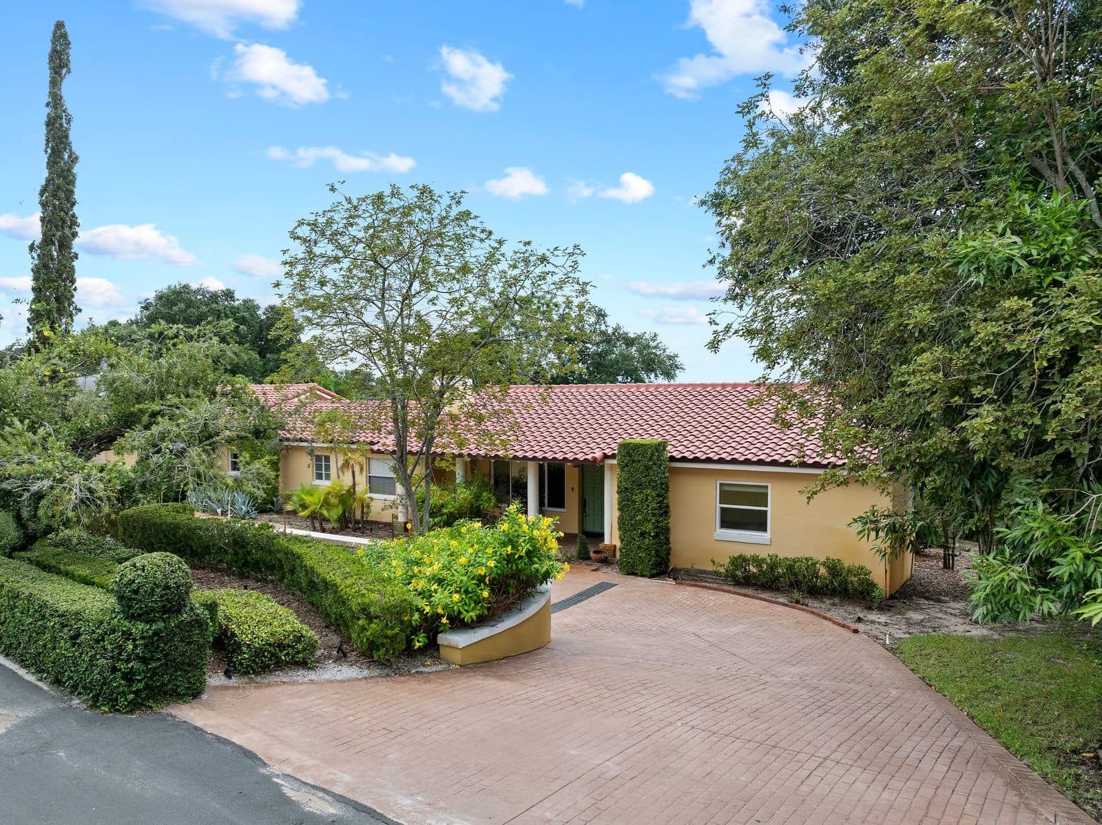 Front of 1121 Sunset Dr, a single-story yellow stucco home with a red tile roof, a columned front porch, and a curved brick driveway lined with hedges