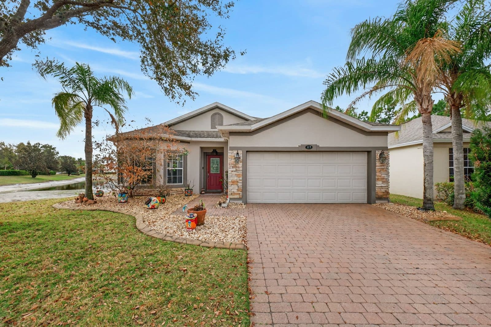 Front of 117 Preston Ave, a single-story home with a red front door, stone accents, a two-car garage, and palm trees, with the pond on the left