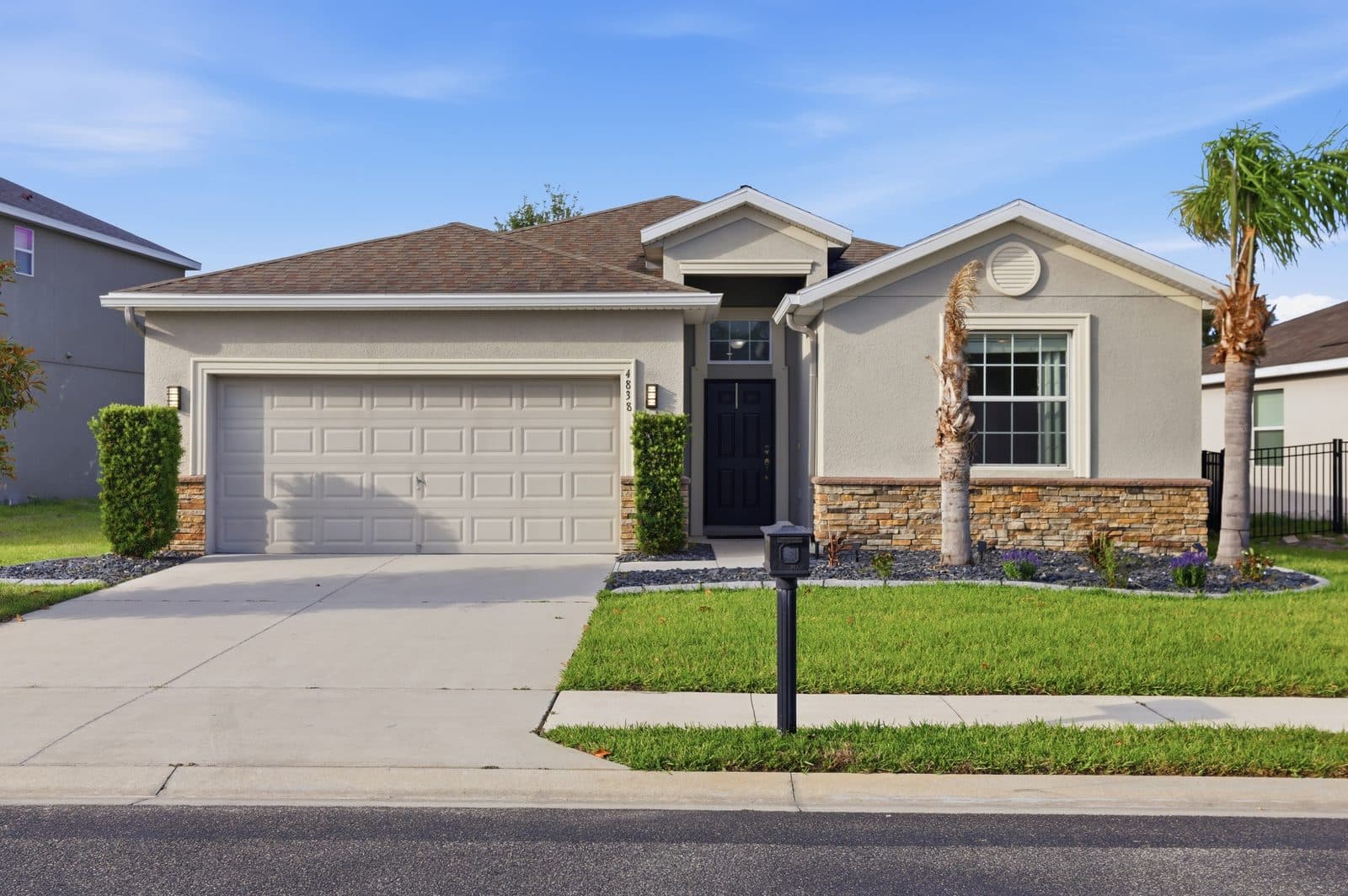 Front of 4838 Cortland Dr, a single-story stucco home with a two-car garage, stone accents, a black front door, and palm trees