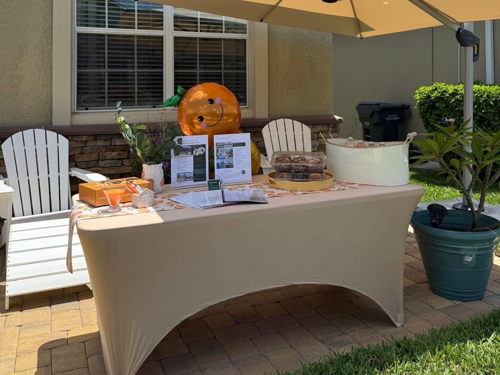 An open house welcome table on the front patio of 3698 Cortland Dr, with listing and Providence flyers, baked goods, bottled drinks, and an orange-shaped balloon under a patio umbrella
