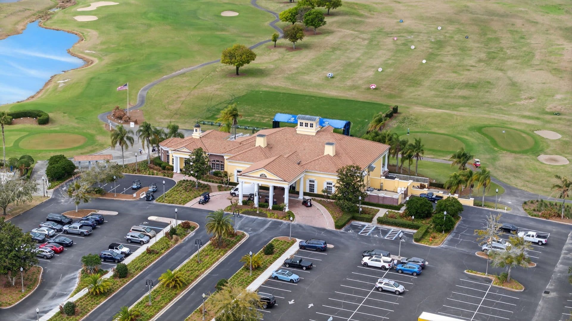 Aerial view of the yellow golf clubhouse in Providence, with its tile roof and columned entry, and putting greens, a driving range, and a lake behind it